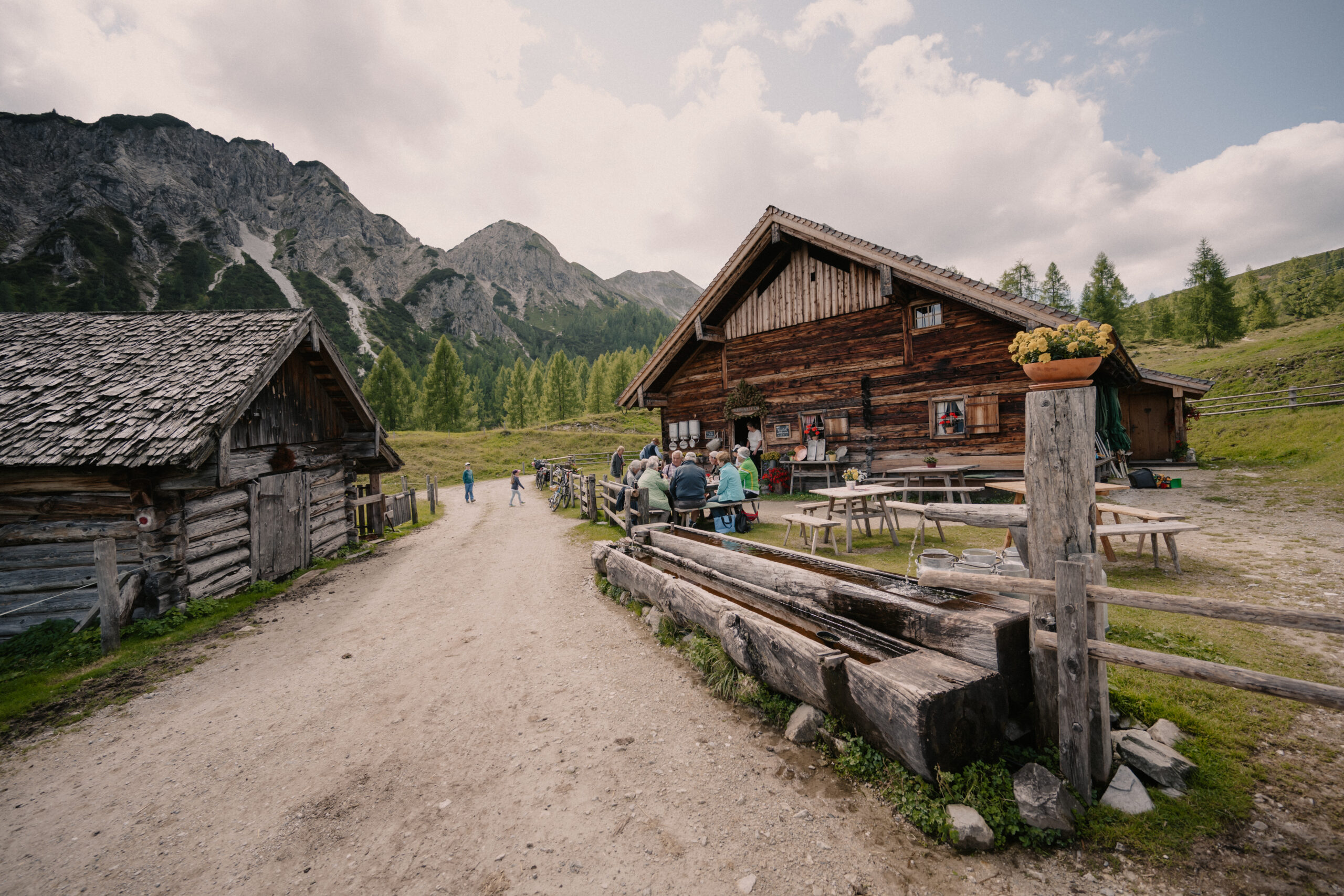 Menschen sitzen an Tischen im Freien bei rustikalen Holzhütten in einem Bergtal mit Bäumen und bewölktem Himmel.