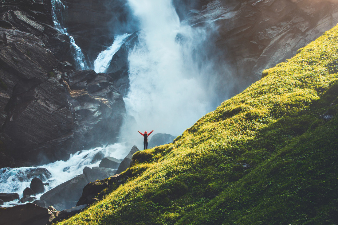 Eine Person mit erhobenen Armen steht auf Felsen in der Nähe eines Wasserfalls, neben einem steilen, sonnenbeschienenen grünen Hang.