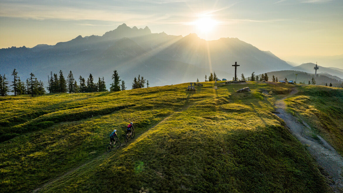 Zwei Radfahrer fahren bei Sonnenaufgang auf einem grasbewachsenen Hügel, mit Bergen und einem großen Kreuz im Hintergrund.