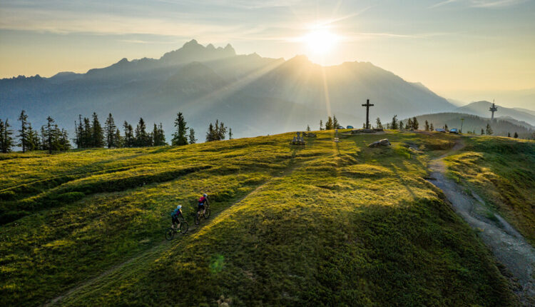 Zwei Radfahrer fahren bei Sonnenaufgang auf einem grasbewachsenen Hügel, mit Bergen und einem großen Kreuz im Hintergrund.