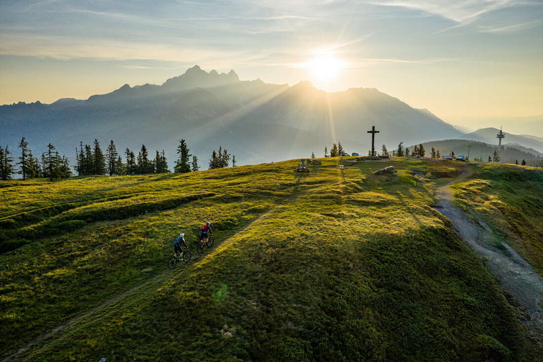Zwei Radfahrer fahren bei Sonnenaufgang auf einem grasbewachsenen Hügel, mit Bergen und einem großen Kreuz im Hintergrund.