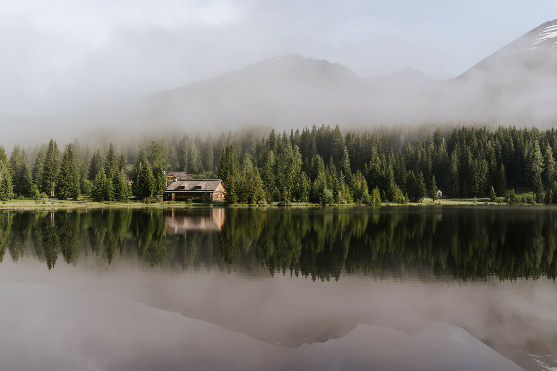 Eine Hütte an einem bewaldeten See, in dessen stillem Wasser sich Bäume und Berge spiegeln, während über dem See Nebel aufsteigt.