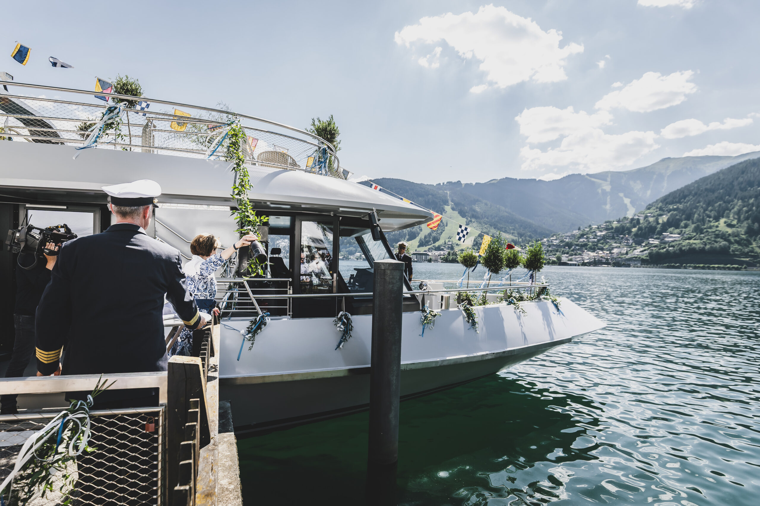 Menschen steigen an einem sonnigen Tag auf einem See, vor dessen Hintergrund sich Berge und eine Stadt abzeichnen, in ein geschmücktes Boot ein.