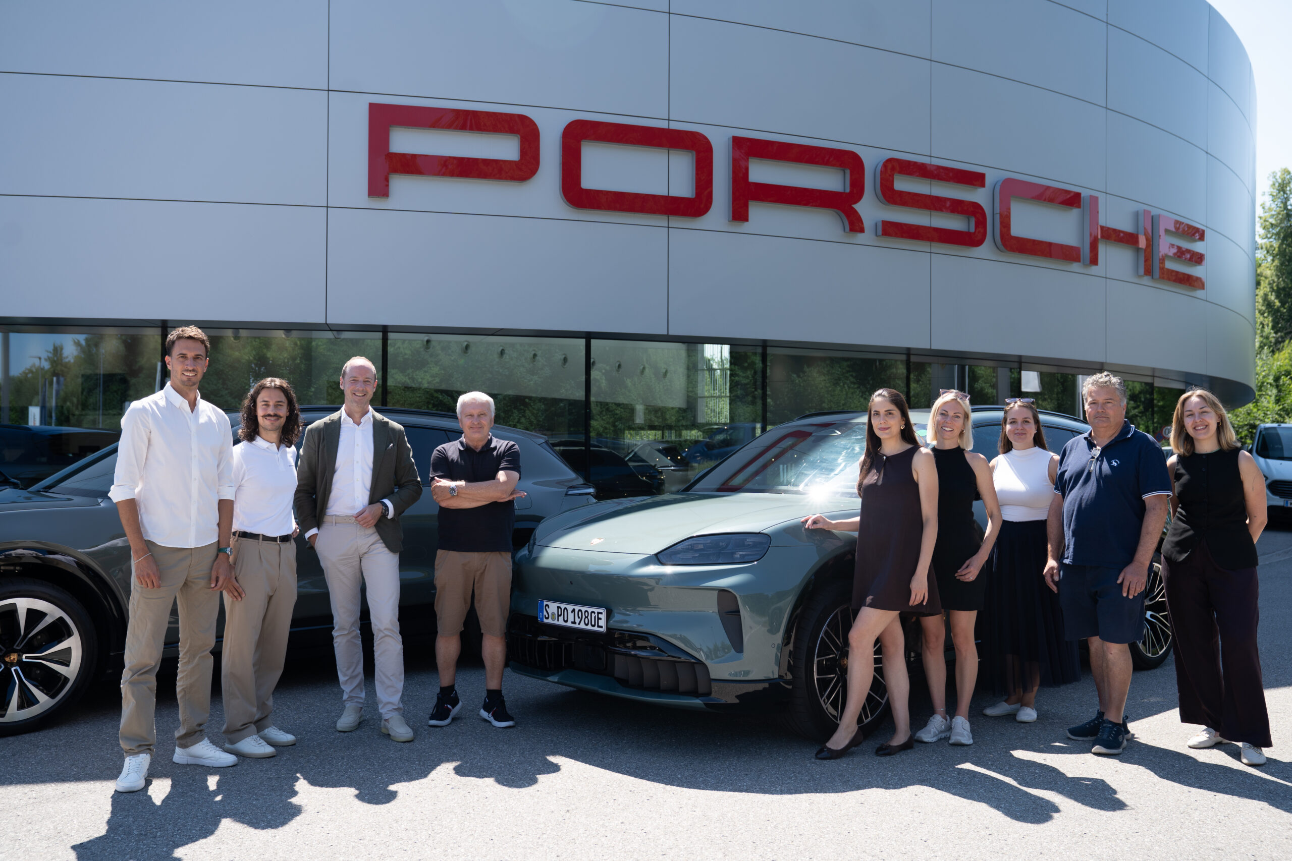 Eine Gruppe von Menschen, die an einem sonnigen Tag vor einem Porsche-Autohaus und einem Porsche-Sportwagen stehen.