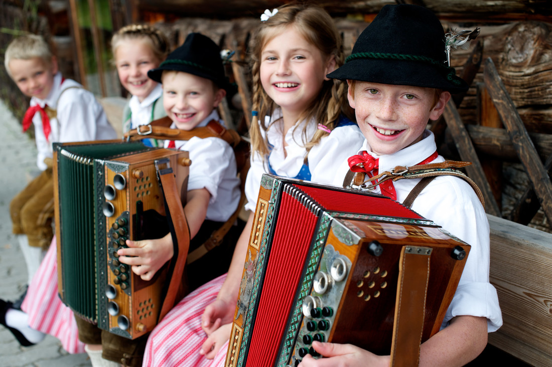 Lächelnde Kinder in traditioneller österreichischer Tracht sitzen auf einer Bank und spielen Akkordeon.