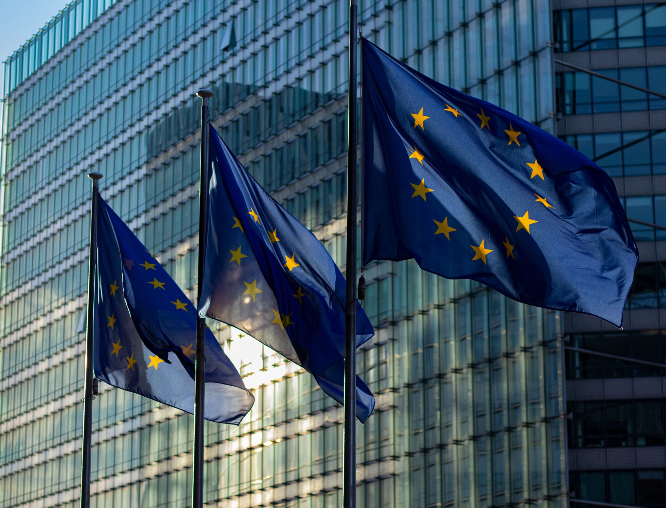 Drei Flaggen der Europäischen Union wehen vor einem modernen Glasgebäude.