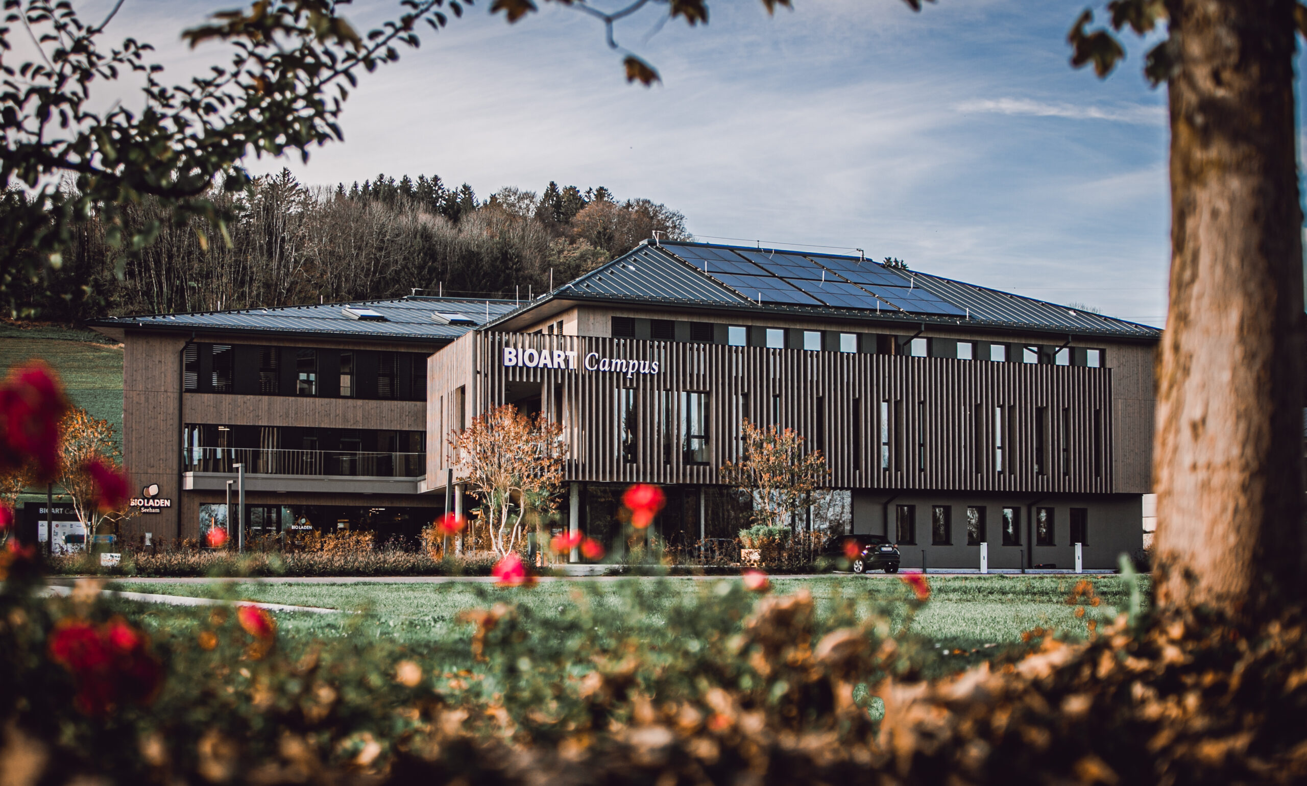 Modernes Gebäude mit der Aufschrift „BIOART Campus“ im Bio-Dorf Seeham, umgeben von Grünflächen, roten Blumen und Bäumen.