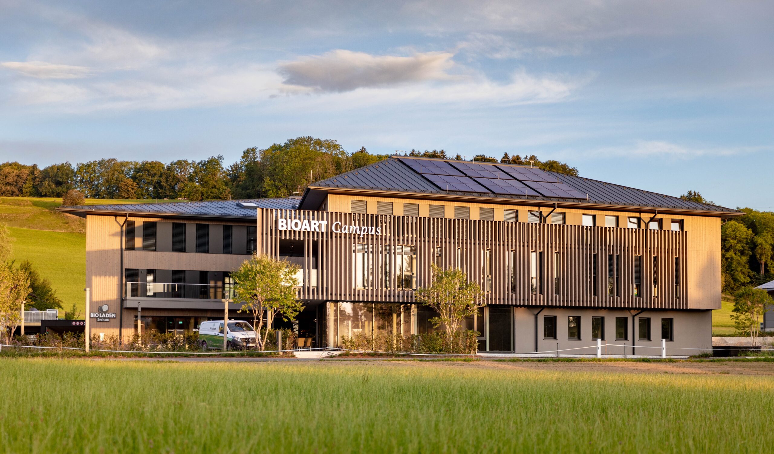 Ein modernes dreistöckiges Gebäude mit der Aufschrift „BioArt Campus“, umgeben von Solarmodulen, Bäumen und grünen Wiesen.