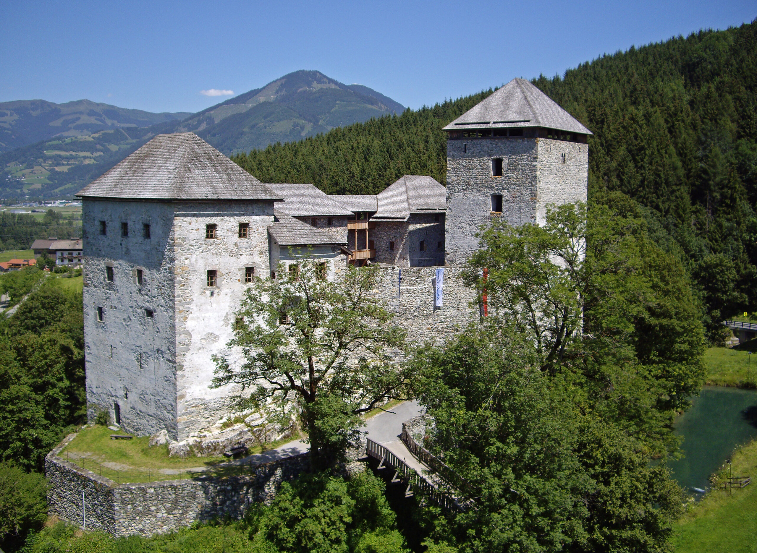 Eine Burg aus Stein mit zwei Türmen, umgeben von Bäumen und Bergen unter einem strahlend blauen Himmel.