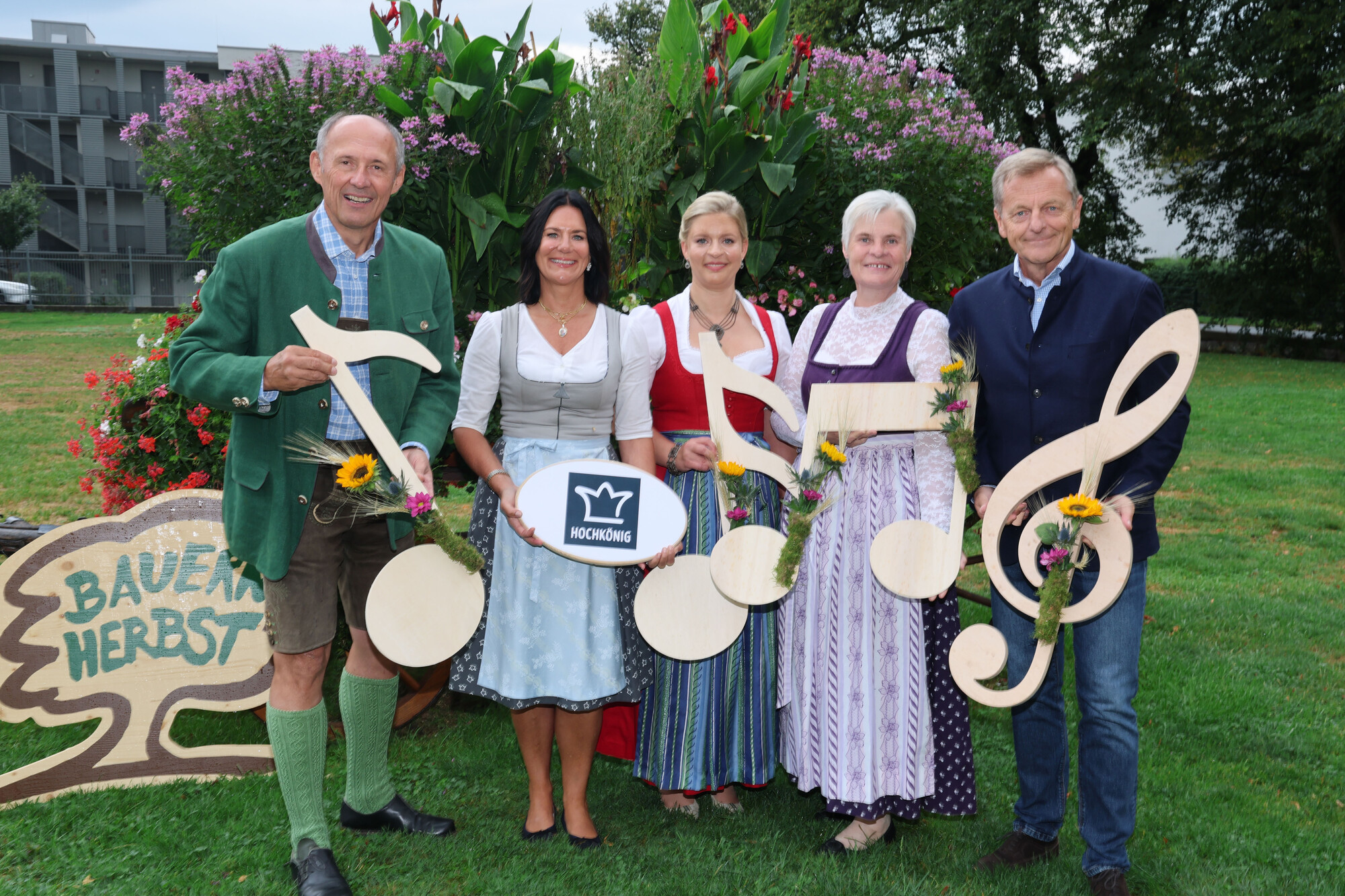 Fünf Personen in traditioneller Tracht halten Noten und Blumen in den Händen und stehen auf einer Wiese neben einem Schild mit der Aufschrift „Bauernherbst“.