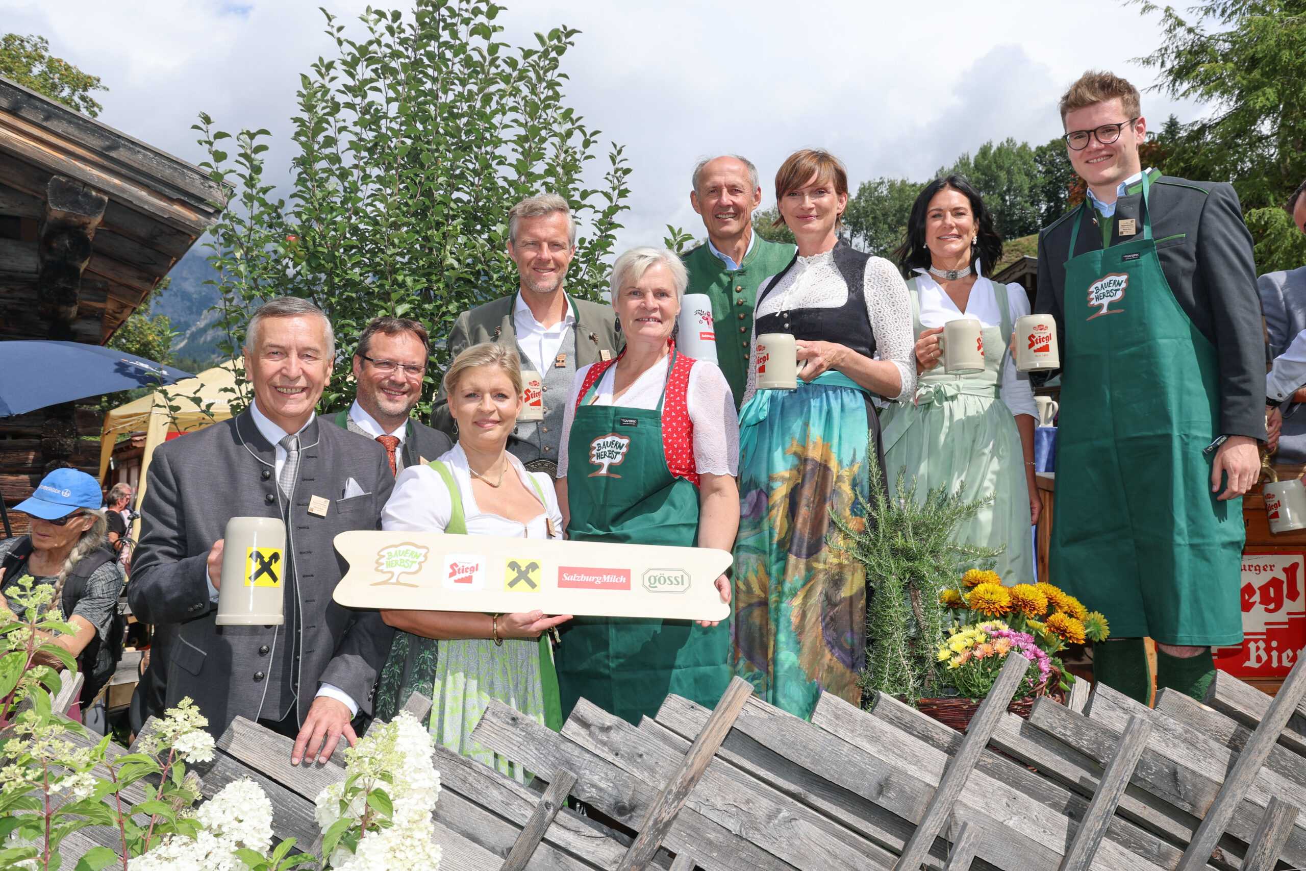 Eine Gruppe in traditioneller österreichischer Tracht steht im Freien, lächelt und hält Bierkrüge und ein Holzschild in den Händen.
