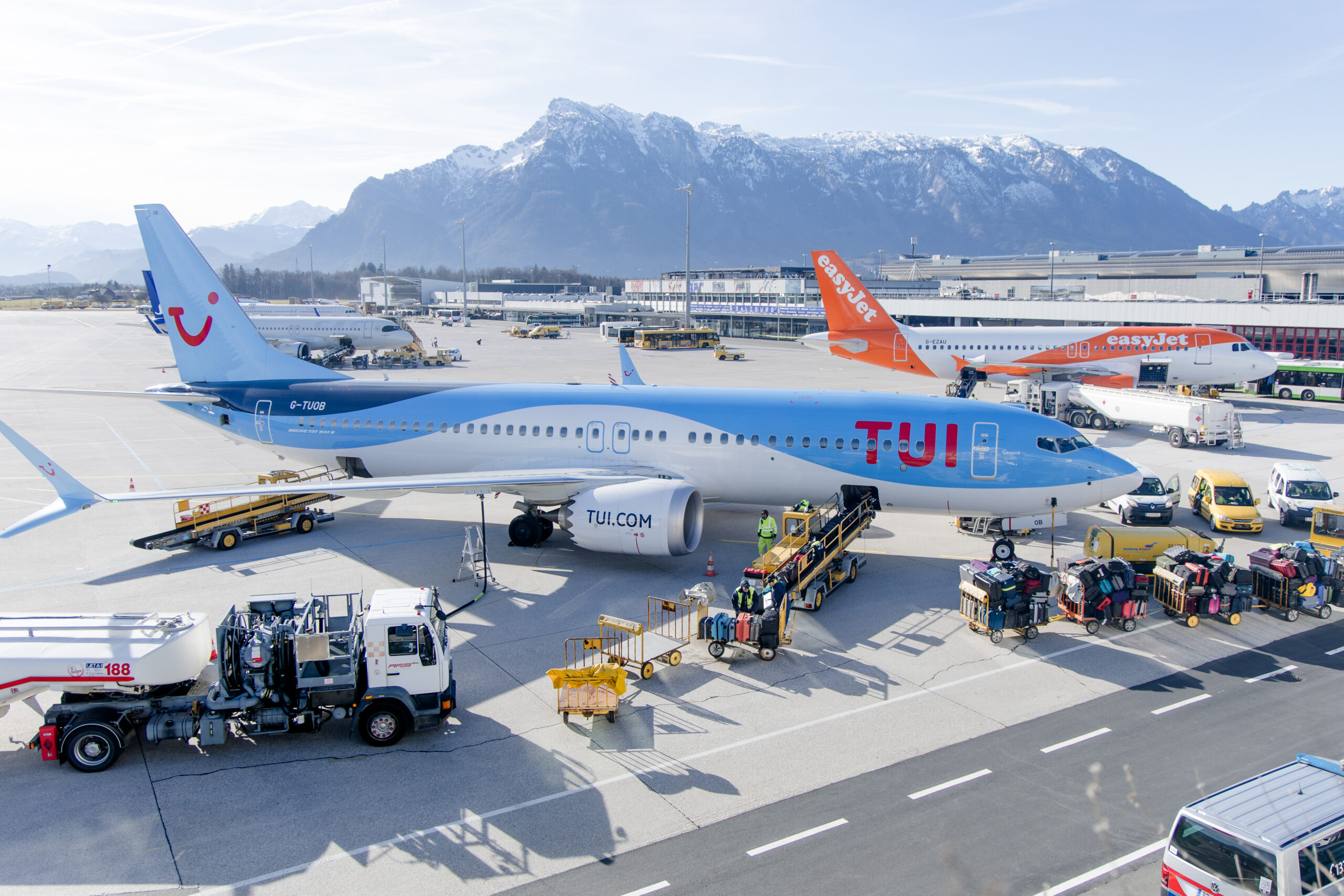 Ein TUI-Flugzeug auf dem Rollfeld des Flughafens, Gepäck wird verladen, im Hintergrund sind Berge und andere Flugzeuge zu sehen.
