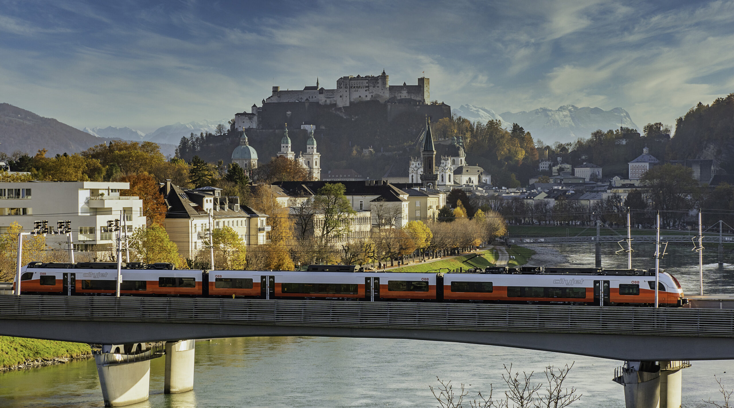 Ein roter Zug überquert eine Brücke in Salzburg, Österreich – die perfekte Kulisse für eine Umfrage zum „Guest Mobility Ticket“.