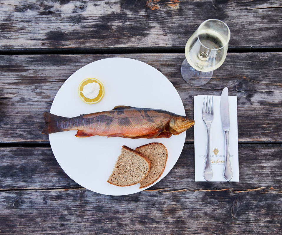 Ein ganzer geräucherter Fisch mit zwei Scheiben Brot, Zitrone und einem Glas Weißwein auf einem rustikalen Holztisch.
