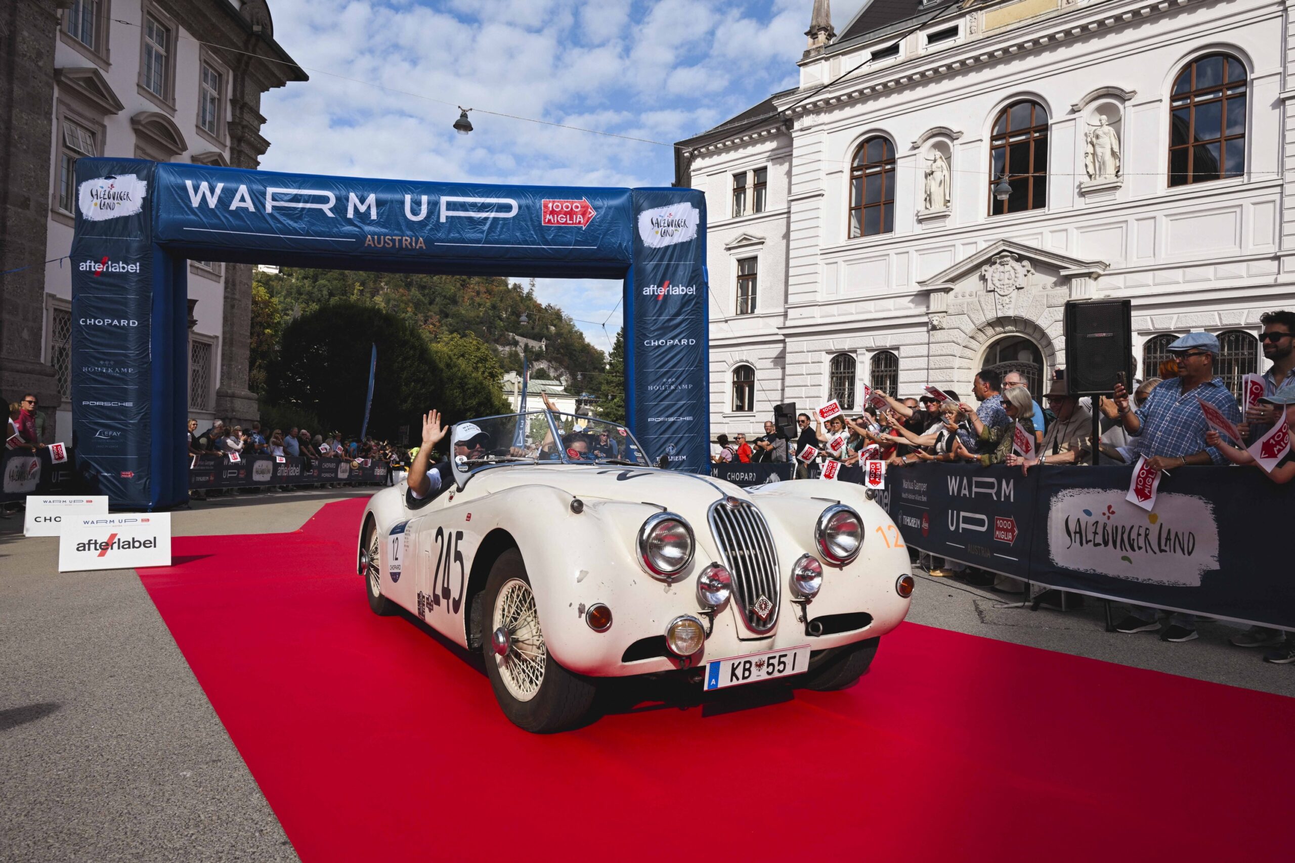Ein klassischer weißer Wagen fährt bei einer Rallye über einen roten Teppich, im Hintergrund jubelt und winkt die Menge.