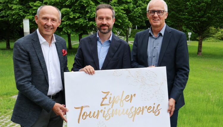 Drei Männer in Anzügen halten auf einer Rasenfläche mit Bäumen in der Nähe ein Schild mit der Aufschrift „Zipfer Tourismuspreis“ hoch.