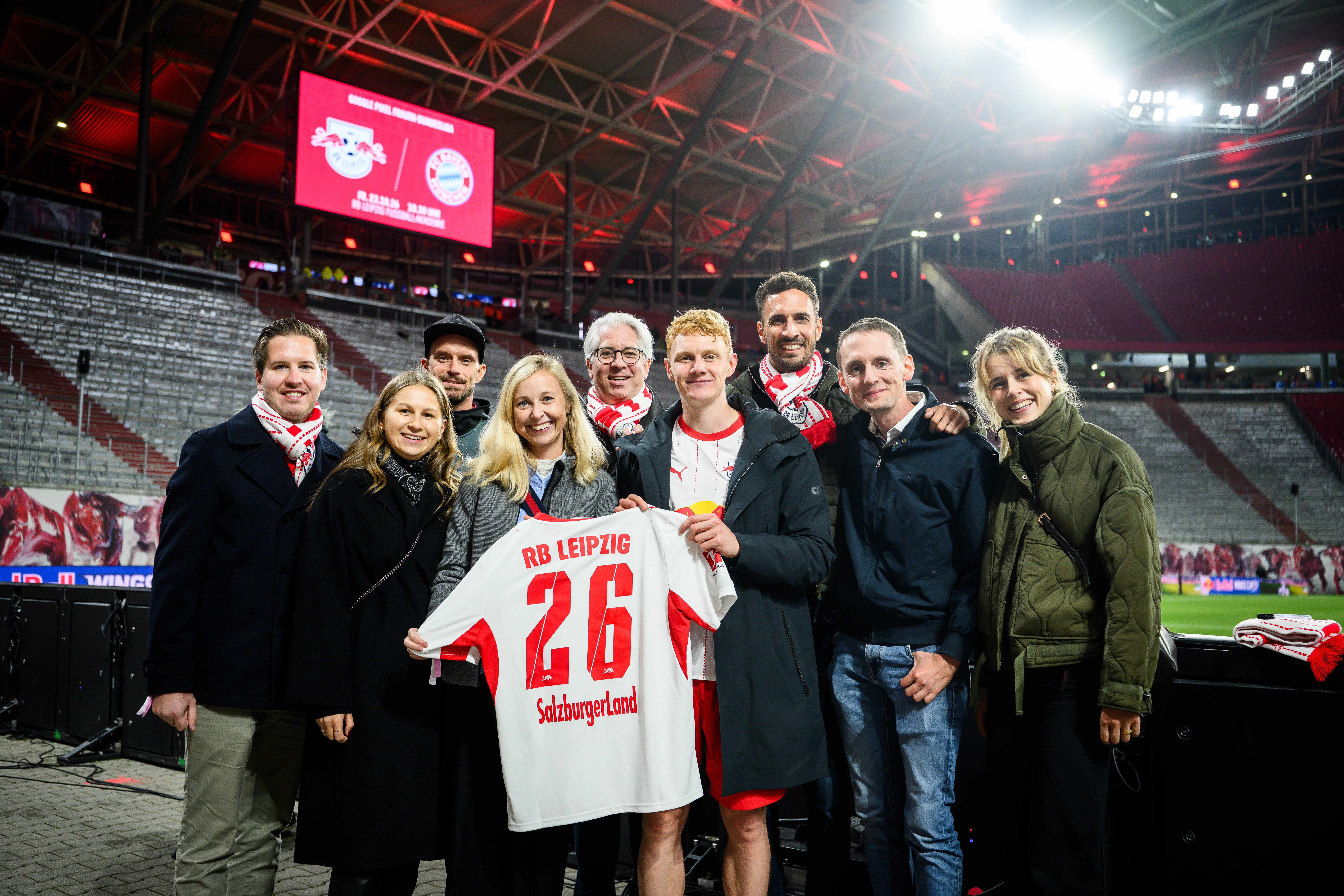 Eine Gruppe von Menschen steht auf einem Fußballplatz und hält ein weißes Trikot mit der Nummer 26 von RB Leipzig auf dem Rücken hoch.